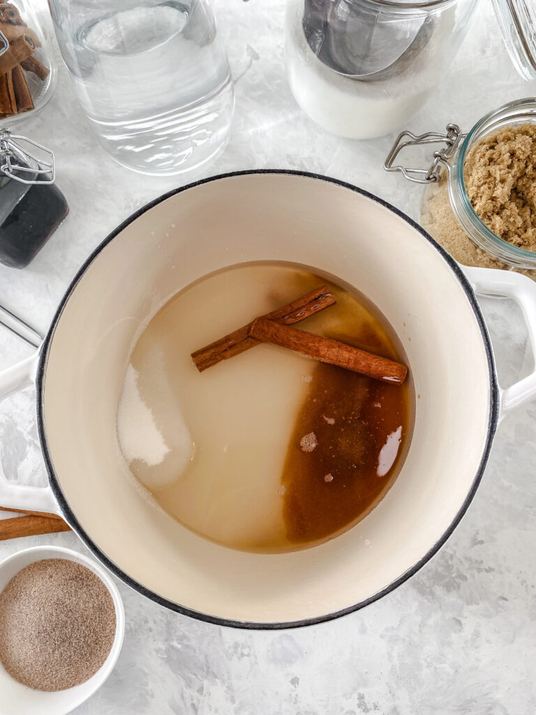 Cinnamon simple syrup ingredients (water, granulated sugar, brown sugar, cinnamon sticks, and vanilla) in a white cast iron pot.