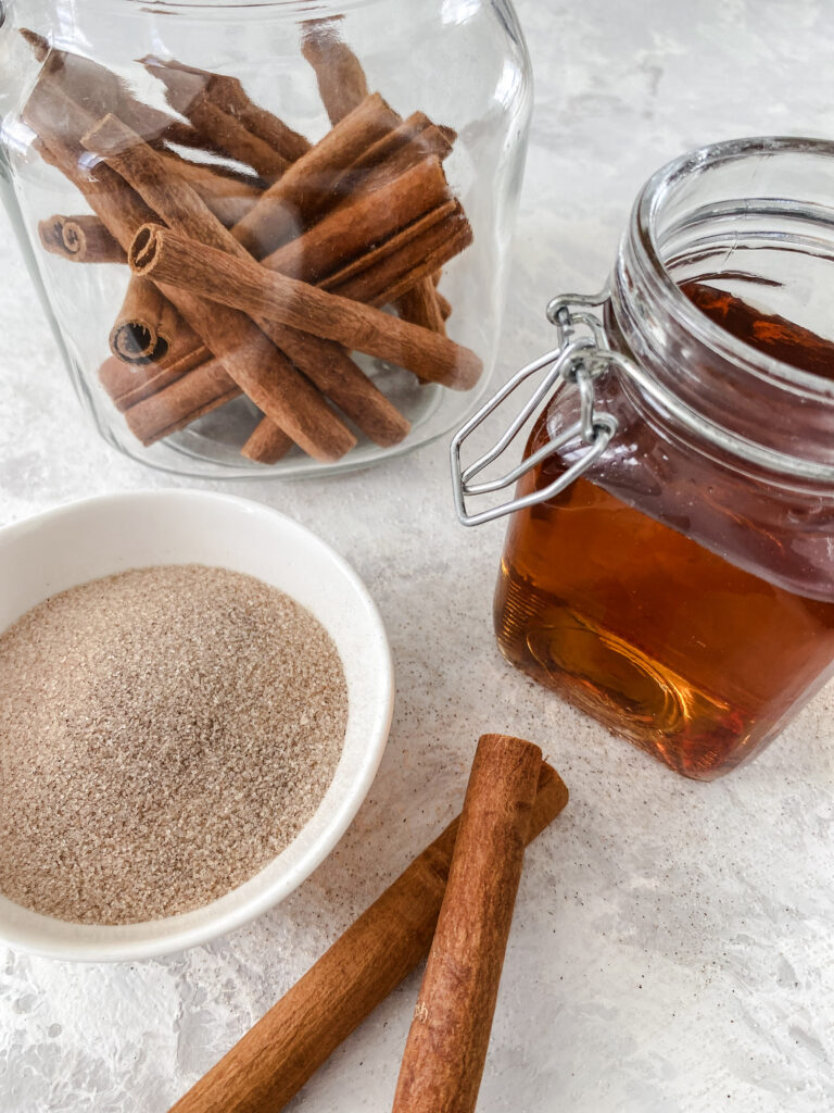 Copycat Starbucks Cinnamon Dolce Syrup in a glass jar beside a bowl of cinnamon sugar and jar of cinnamon sticks.