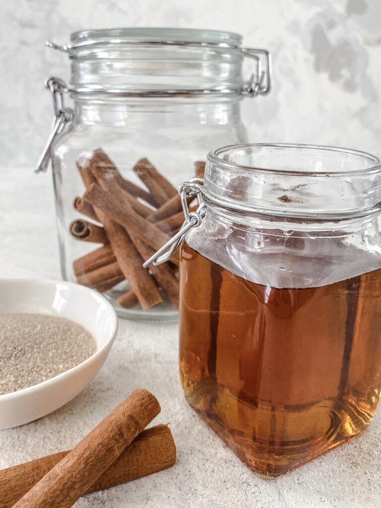 Copycat Starbucks Cinnamon Dolce Syrup in a glass jar beside a bowl of cinnamon sugar and jar of cinnamon sticks.