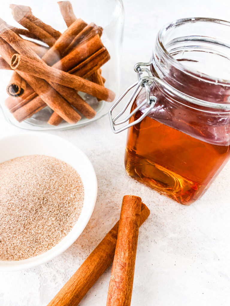 A jar of Copycat Cinnamon Dolce Syrup beside a jar of cinnamon sticks and sparkling cinnamon sugar.