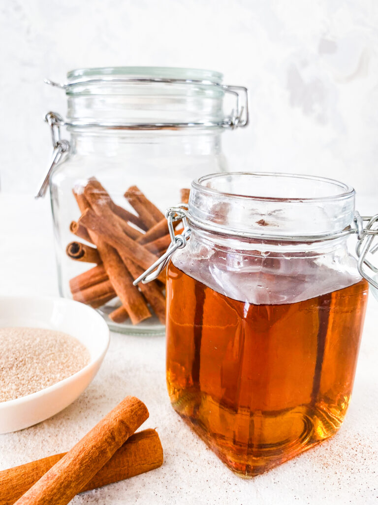 A jar of Copycat Cinnamon Dolce Syrup beside a jar of cinnamon sticks and sparkling cinnamon sugar.