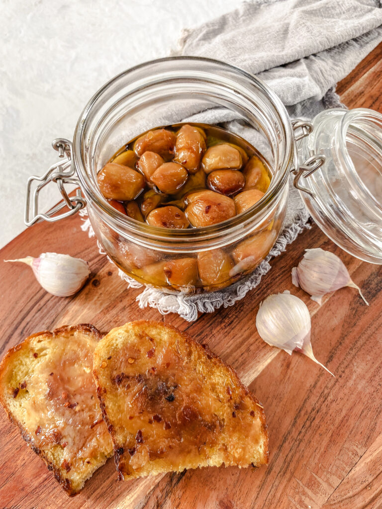 Garlic confit spread on crostini on top of a wooden cutting board, next to a jar of garlic confit.