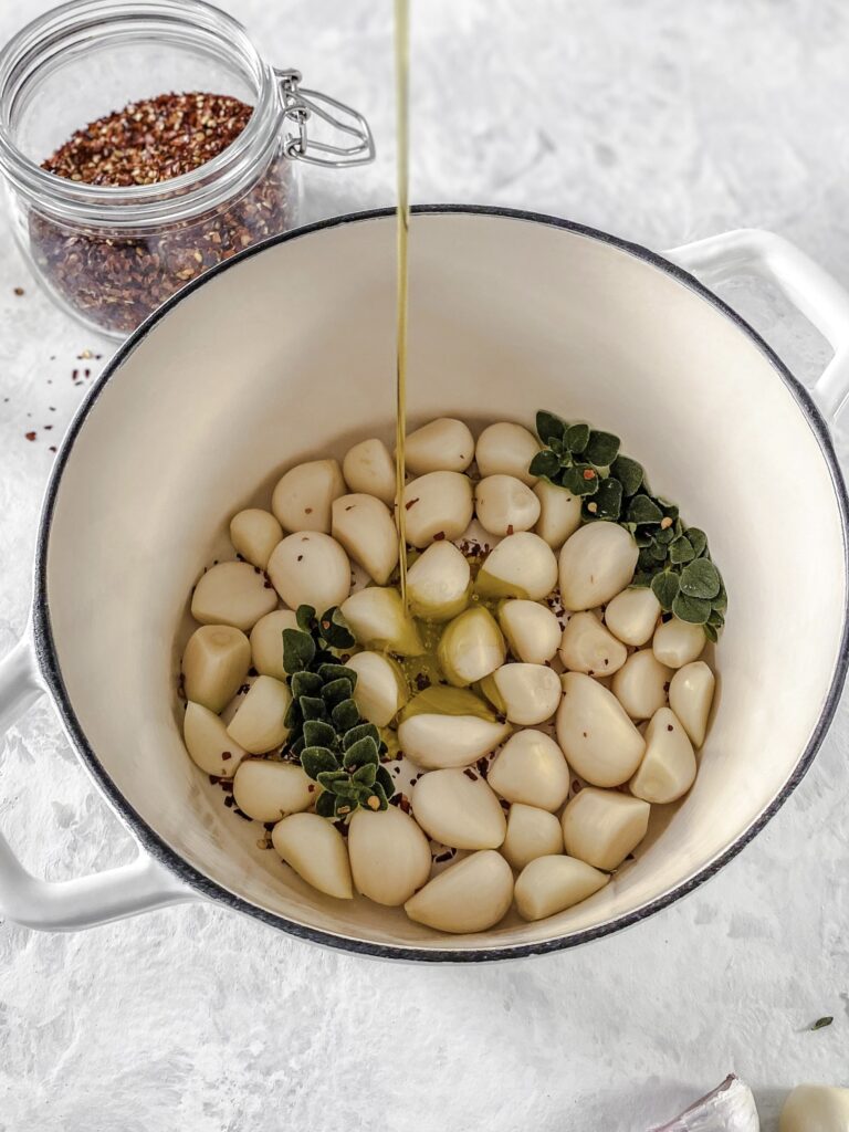 Oil being drizzled over garlic, oregano, and red pepper flakes in a white cast-iron pot.