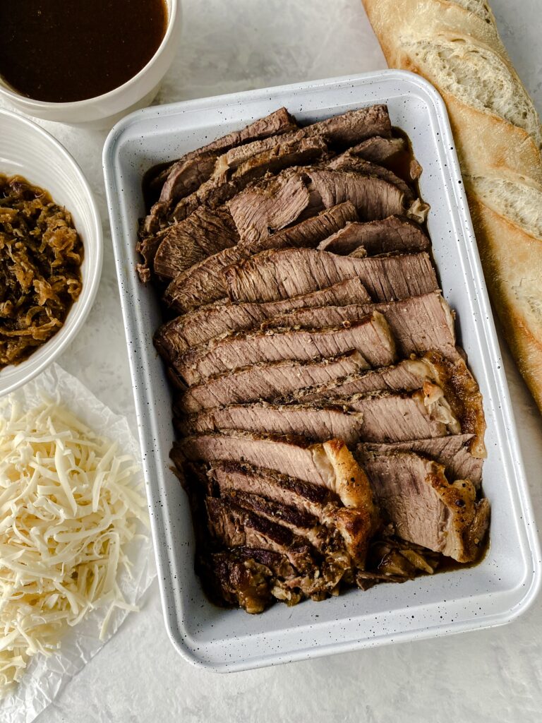 Roast beef in a tray beside a baguette, caramelized onions, shredded cheese, and jus.