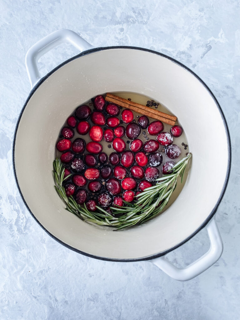 A white cast-iron pot with cranberries, rosemary, peppercorns, sugar, apple cider vinegar, and a cinnamon stick.