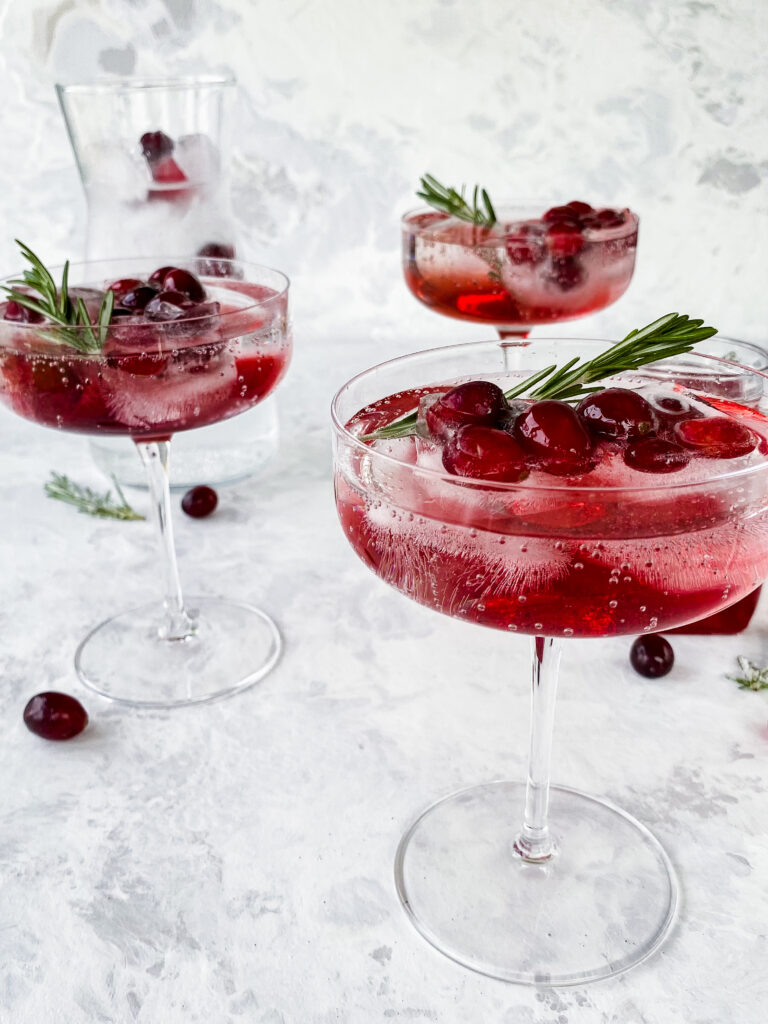 Three coup glasses of cranberry shrub and sparkling water with rosemary and cranberry ice cubes.