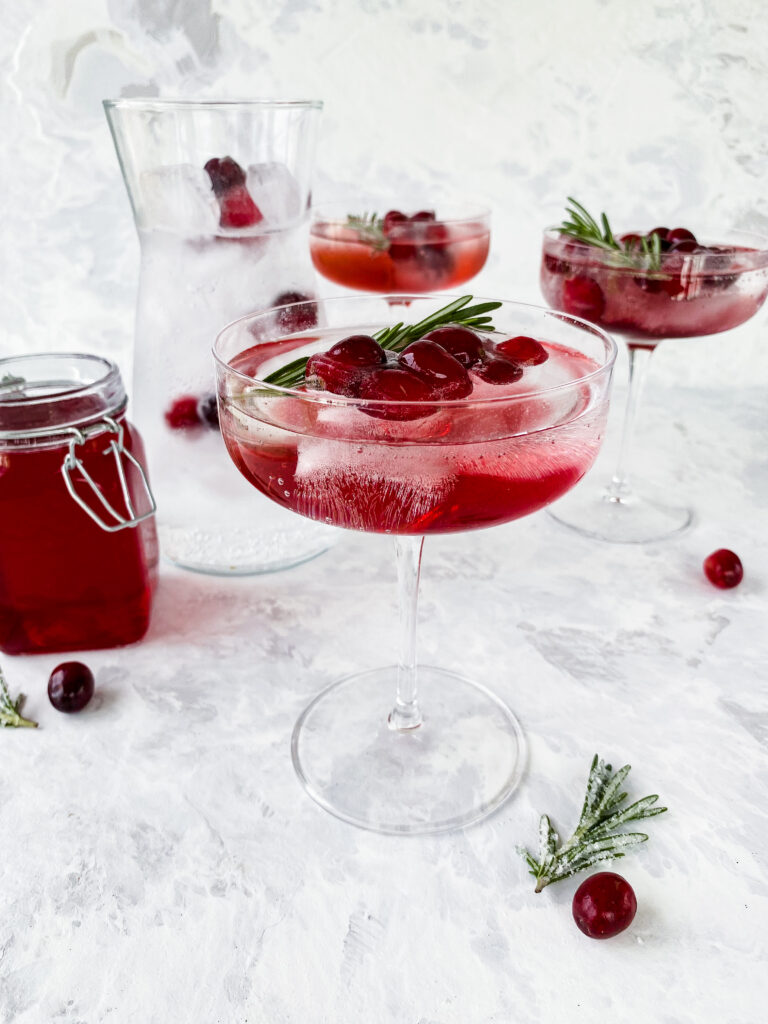 Cranberry shrub and sparkling water in a coup glass with rosemary and cranberry ice cubes.