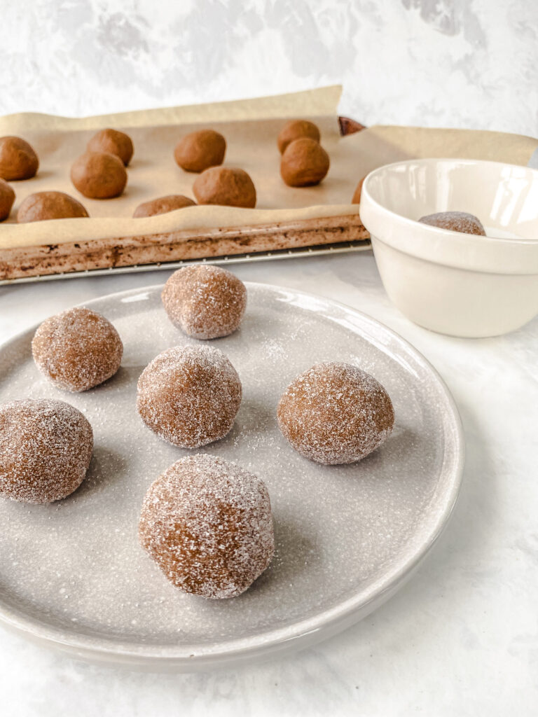 Sugar rolled cookie dough balls on a plate in front of a baking sheet holding more gingerbread cookie dough balls not yet rolled in sugar.