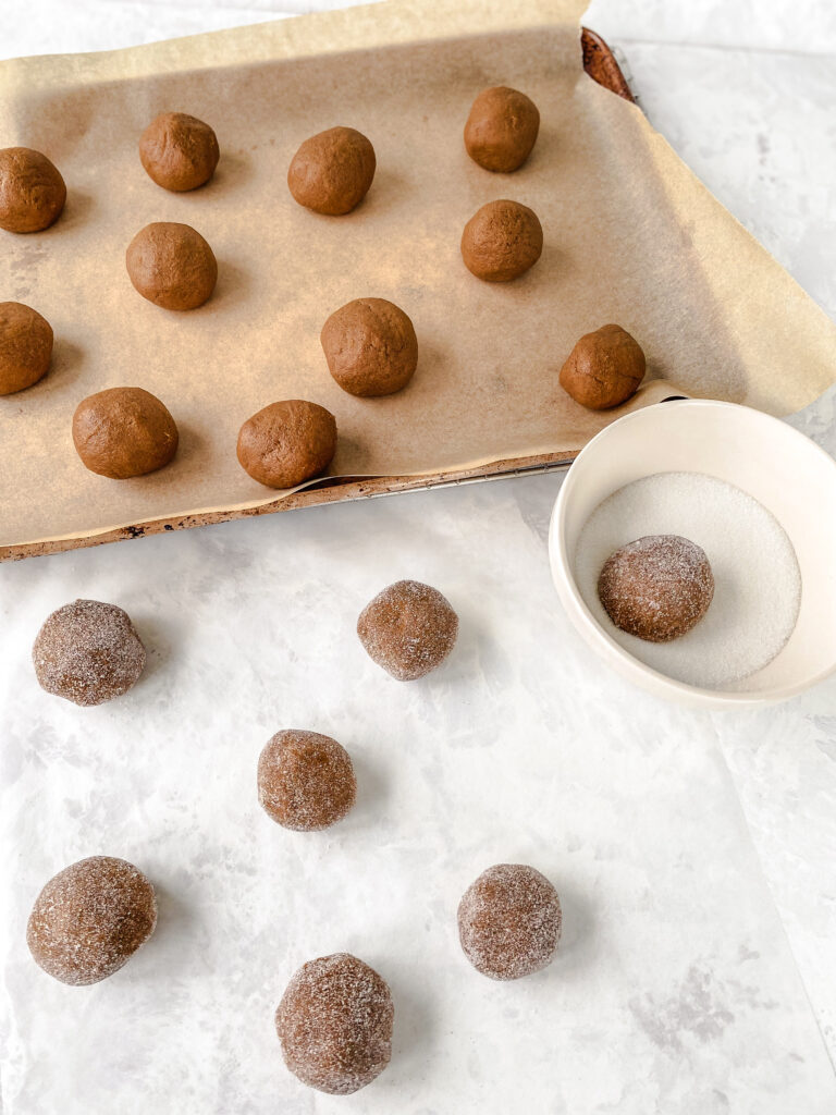 Gingerbread cookie dough balls on a parchment-lined baking sheet next to a bowl of sugar and sugar rolled cookie dough balls.