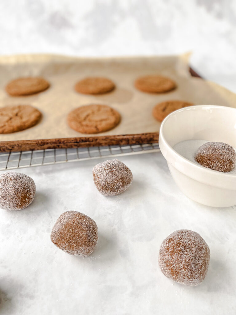 Ginger molasses cookies cooling on a baking sheet behind a bowl of sugar and sugar rolled cookie dough balls.