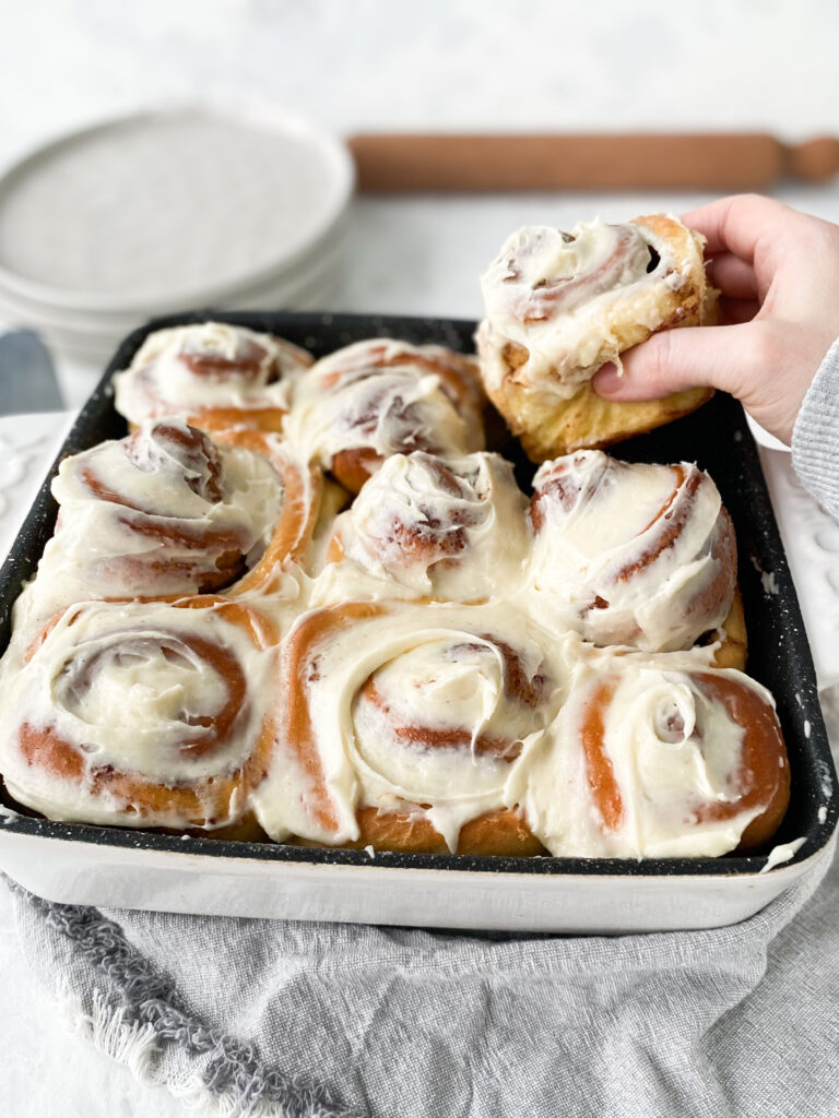 A hand grabbing a cinnamon bun topped with cream cheese frosting.