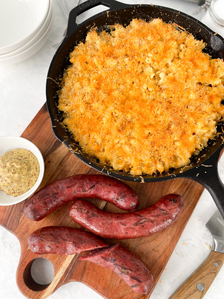 Baked mac and cheese in a cast-iron skillet beside smoked sausages and dijon mustard.