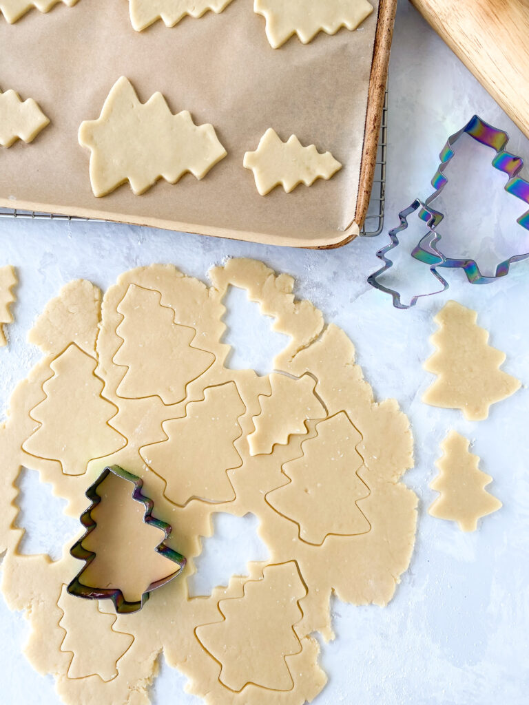 Sugar cookie dough being cut out into evergreen trees and placed on a baking sheet.