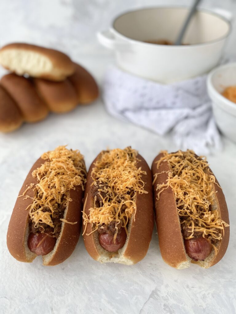 Three hot dogs topped with Cincinnati chili and shredded cheddar cheese (aka Cincinnati Chili Dogs).