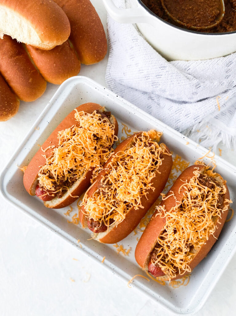 Three hot dogs topped with Cincinnati chili and shredded cheddar cheese (aka Cincinnati Chili Dogs).