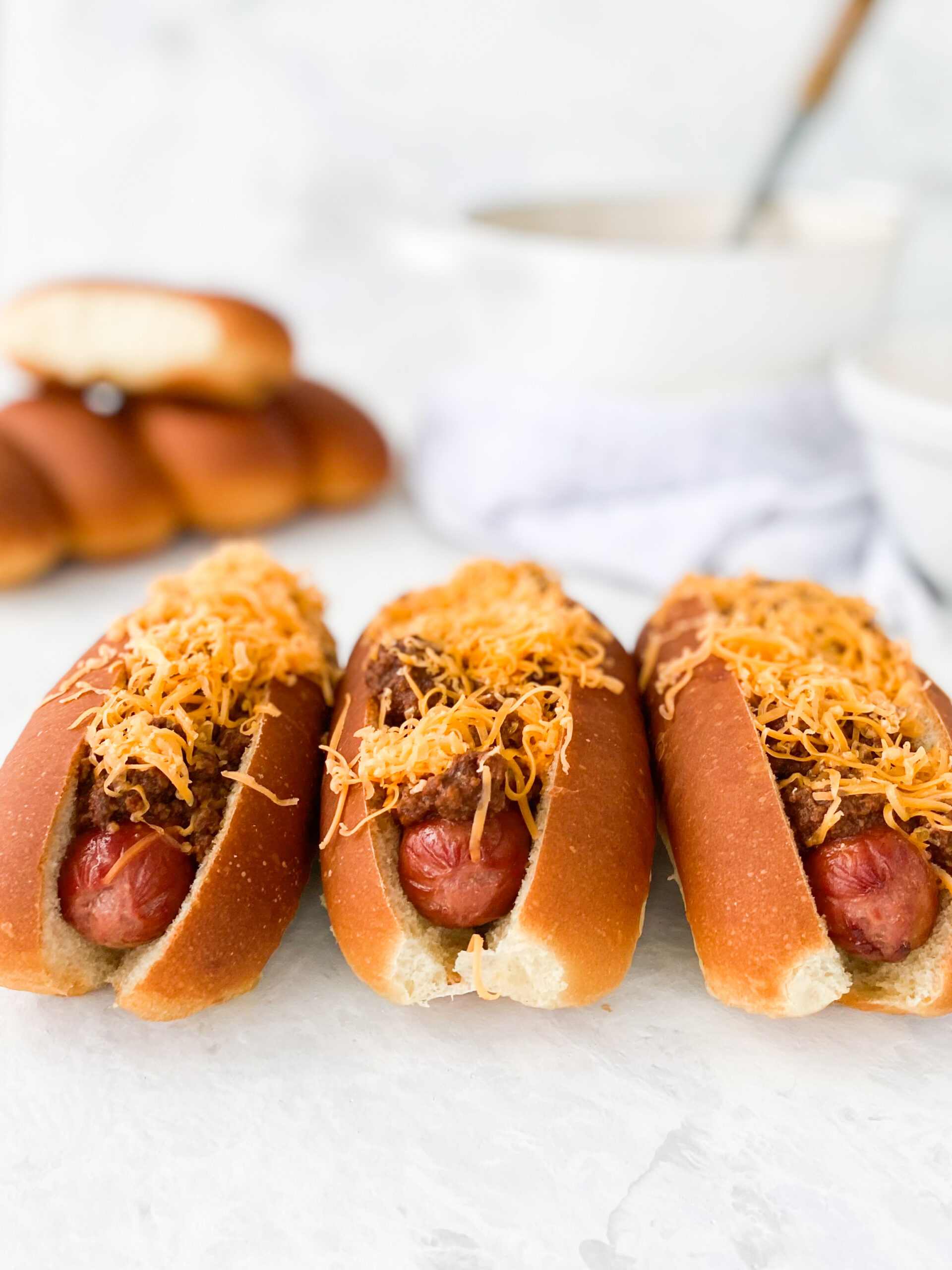 Three hot dogs topped with Cincinnati chili and shredded cheddar cheese (aka Cincinnati Chili Dogs).