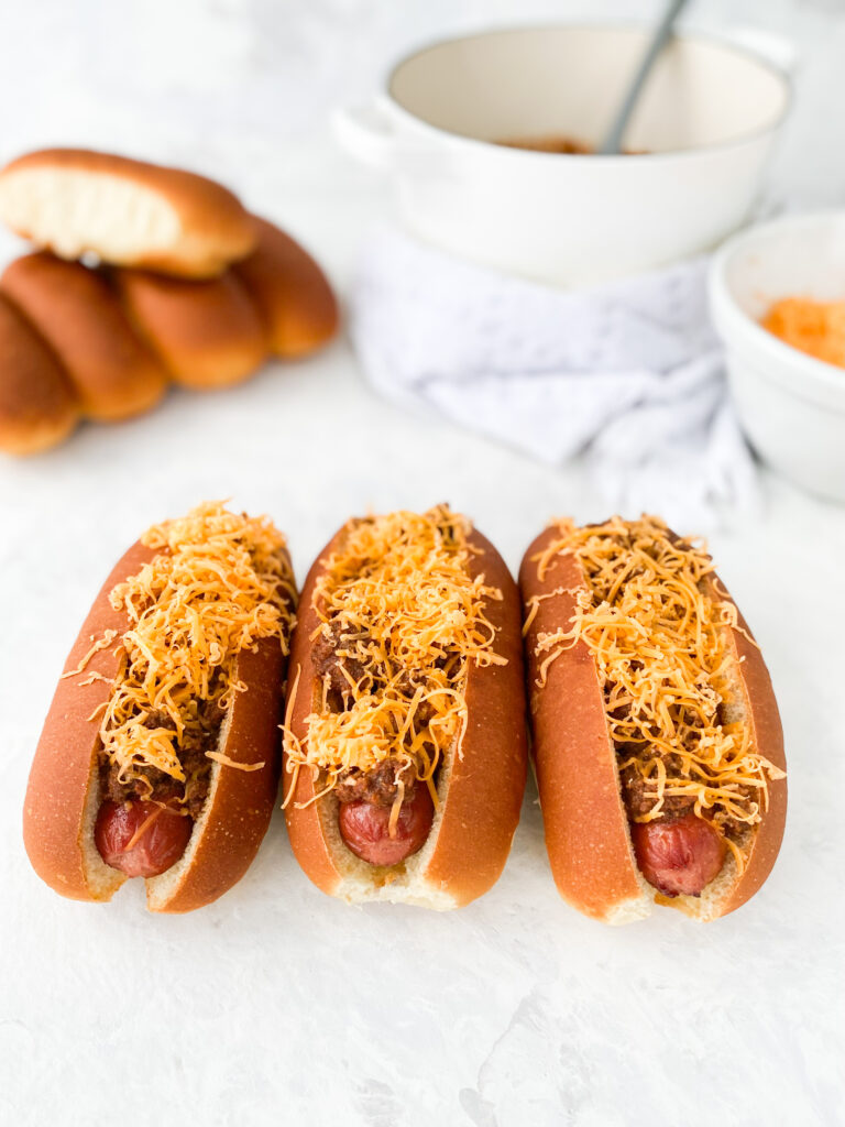 Three hot dogs topped with Cincinnati chili and shredded cheddar cheese (aka Cincinnati Chili Dogs).