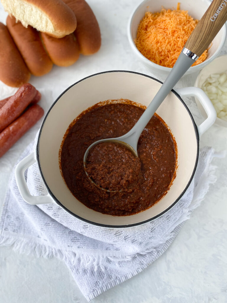 Cincinnati chili in a cast-iron pot next to soft hot dog buns, grilled hot dogs, shredded cheddar cheese, and chopped onions.