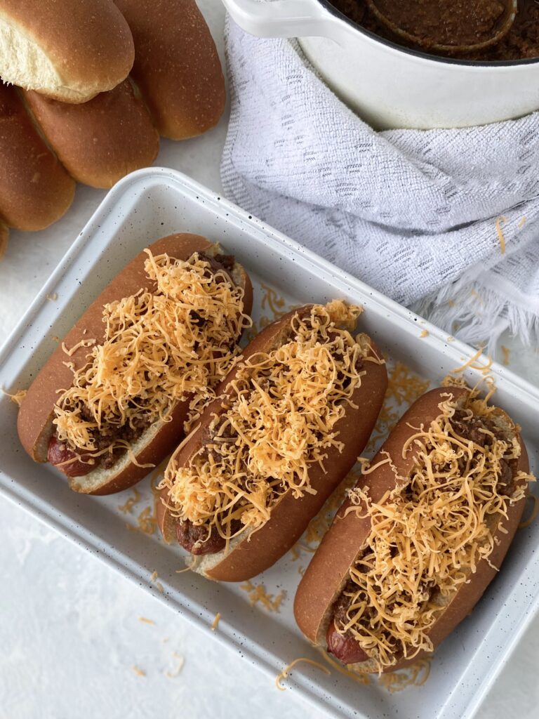 Three hot dogs topped with Cincinnati chili and shredded cheddar cheese (aka Cincinnati Chili Dogs).