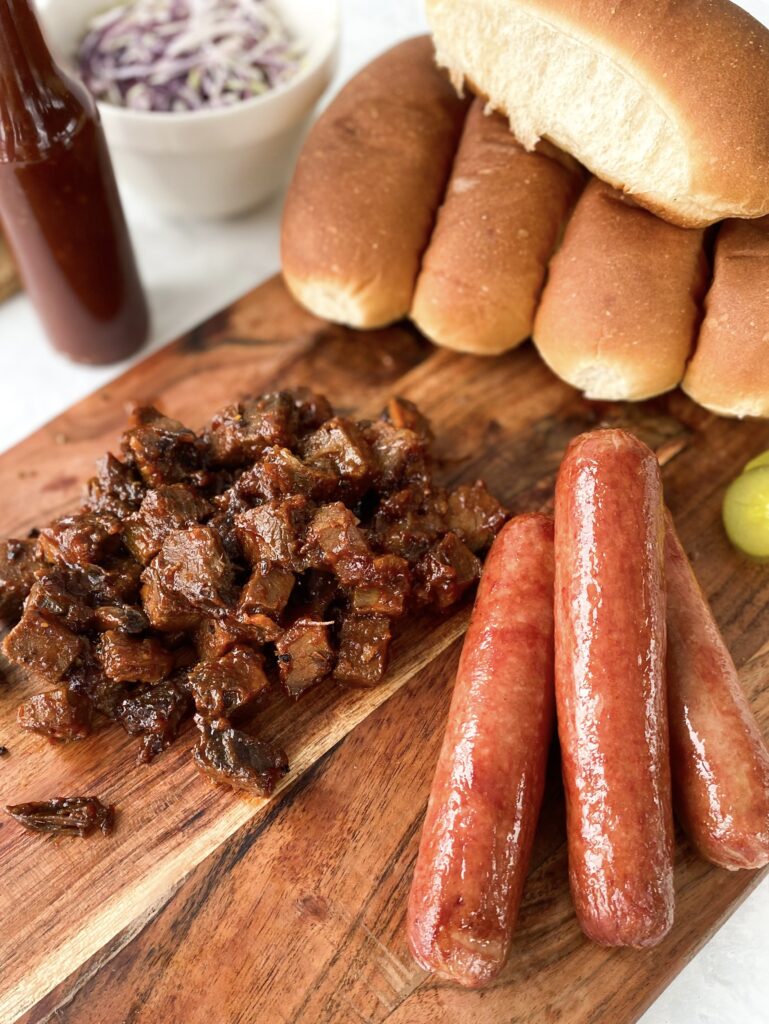 Beef hot dogs, BBQ brisket, hot dog buns, and pickles on a wooden cutting board.