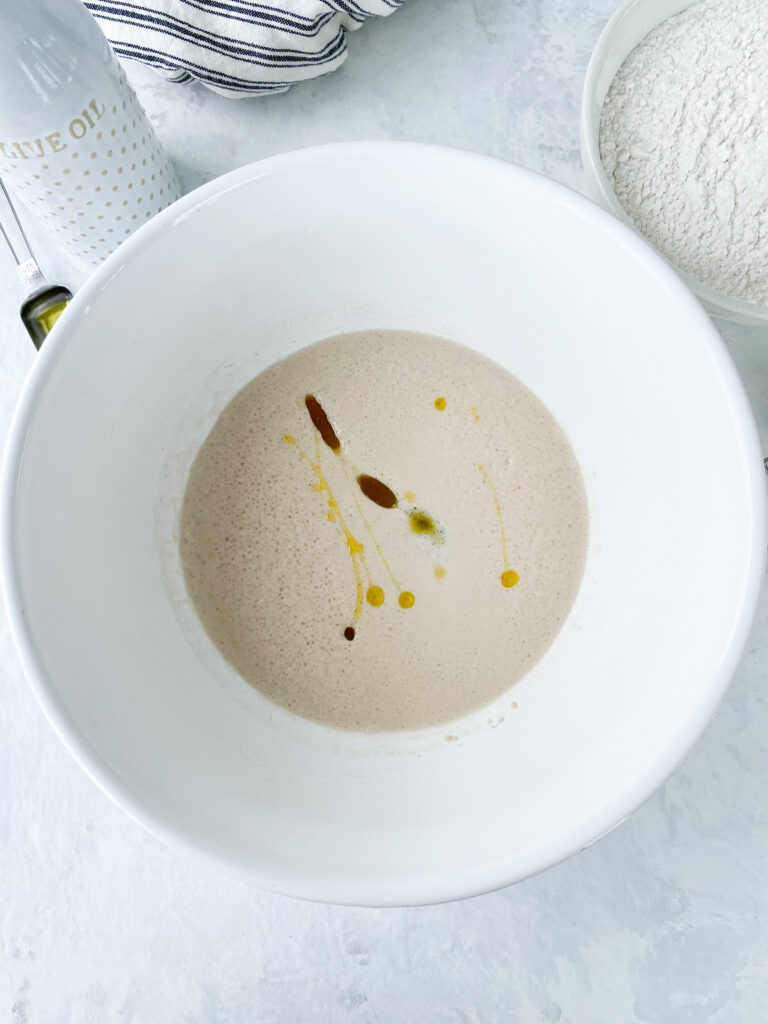 Olive oil added to activated yeast in a white bowl.