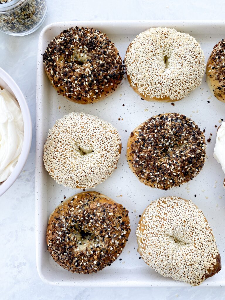 Homemade bagels topped with everything bagel seasoning and sesame seeds with whipped cream cheese on a baking sheet.