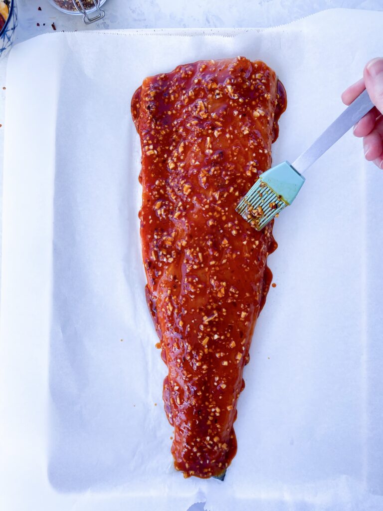 Whole side of salmon on a parchment-lined baking sheet being brushed with fiery garlic chili sauce.