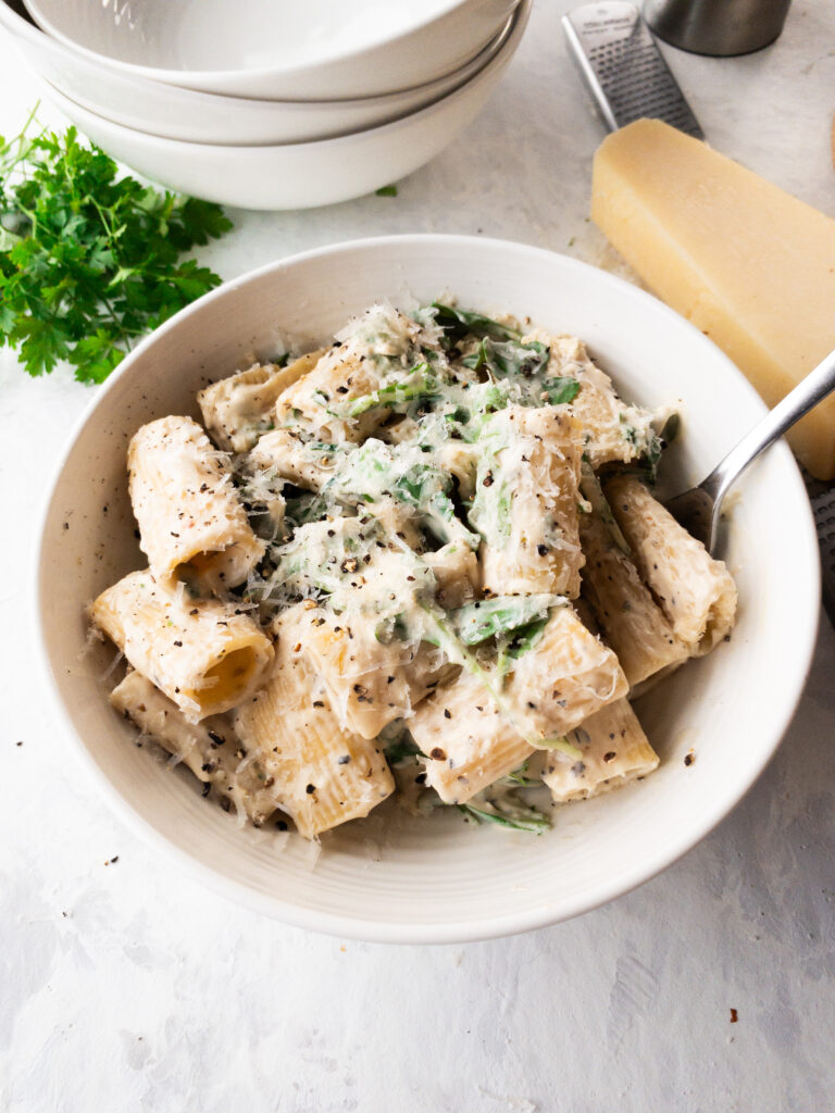 A bowl of rigatoni pasta in a cream sauce (pasta bianca) with arugula, parmesan, and cracked pepper.