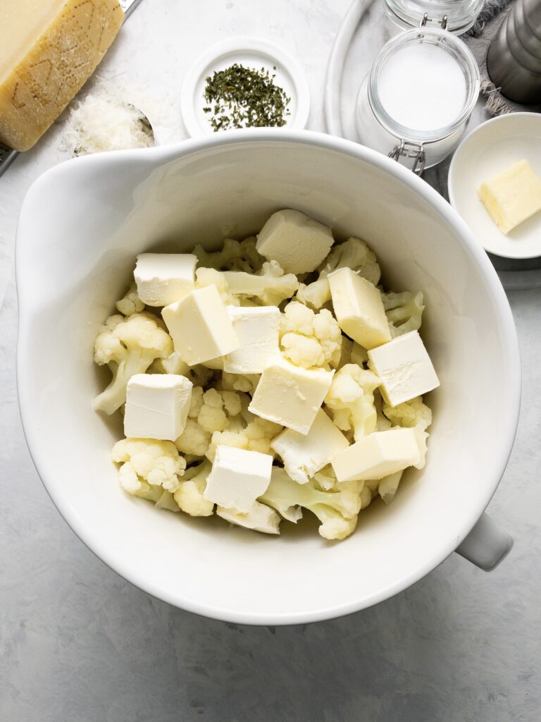 Cooked cauliflower topped with cubes of cream cheese and butter for a creamy, low-carb mashed cauliflower side dish.