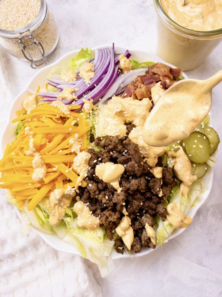 Homemade Mac Sauce spooned over Big Mac Salad (made with iceberg lettuce, seasoned ground beef, shredded cheddar cheese, sliced red onion, crumbled bacon, pickles, and sesame seeds.)